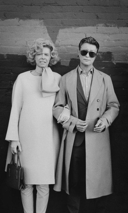 Black and white photo of a couple against a brick wall, one in a cream bow-neck dress, one in a trench coat