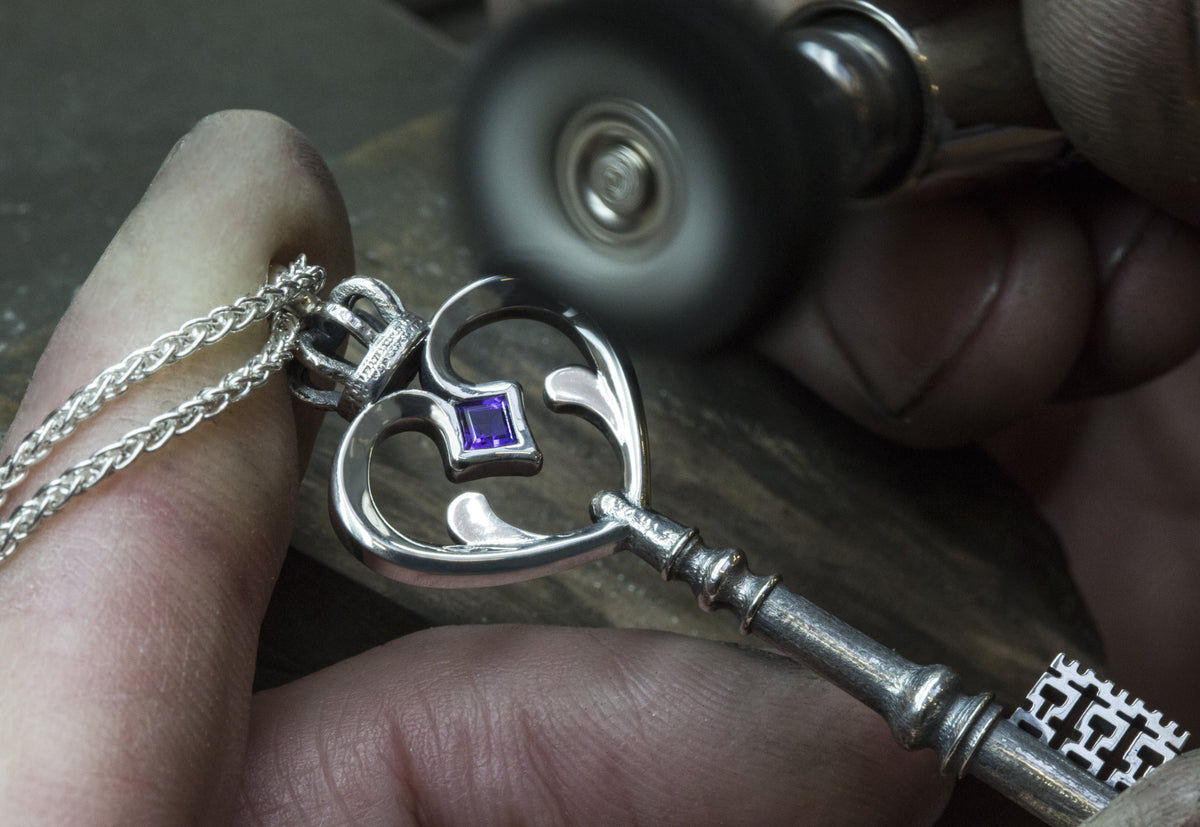 Silver key pendant with a heart-shaped bow, a purple square stone and crown top, being polished by hand