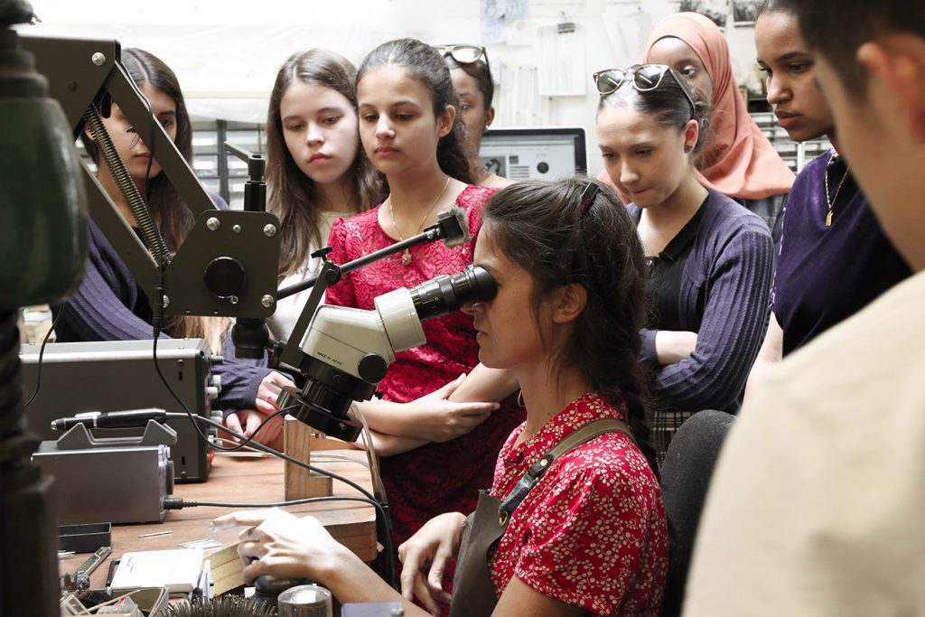 Chelsea Academy students watching one of their group look through a microscope at a jeweller's bench