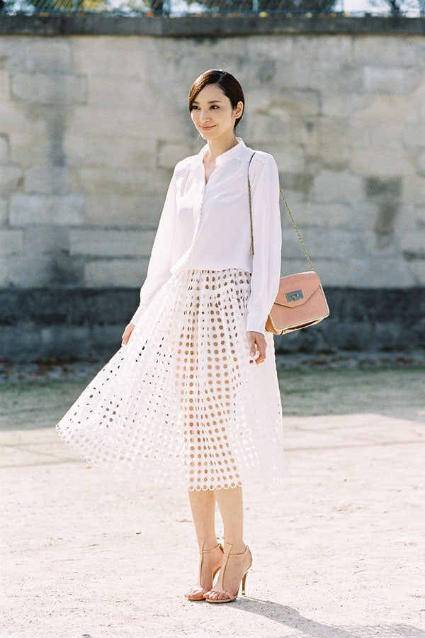 Woman in a white shirt and white cut-out midi skirt with a pink shoulder bag and nude heels by a stone wall