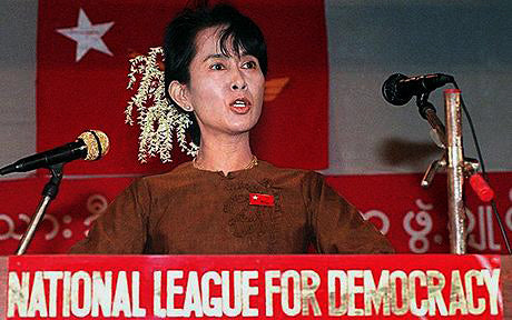 Pro-democracy leader speaking at a National League for Democracy podium in front of a red flag in Burma