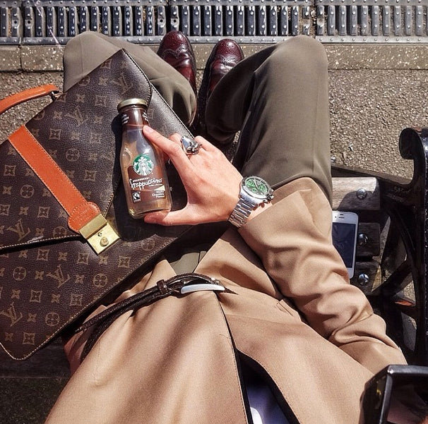 Man seated on a London bench wearing a silver bull ring, steel watch and camel coat, holding a coffee bottle