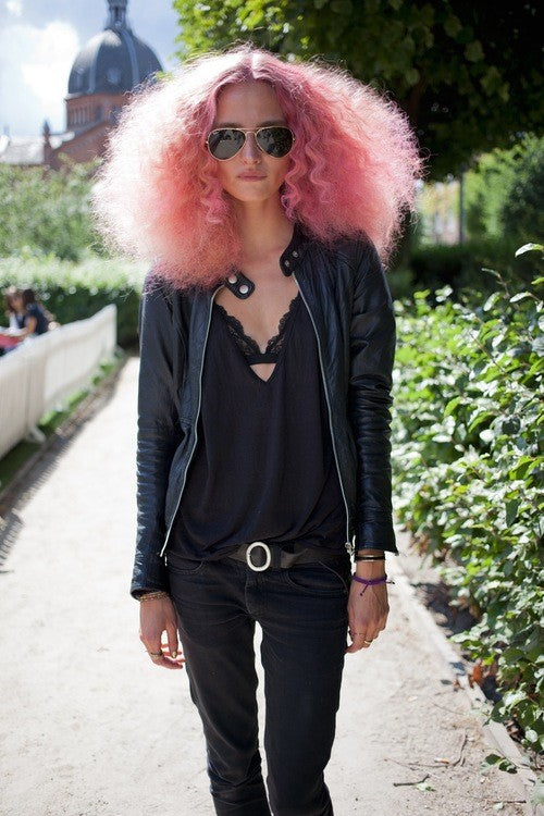 Woman with long pink curly hair and aviator sunglasses in a black leather jacket, wearing bracelets