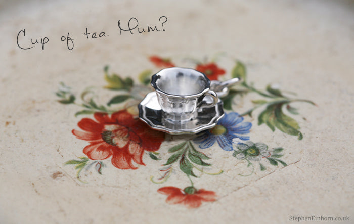 Silver teacup and saucer charm on a floral plate, handwritten Cup of tea Mum?