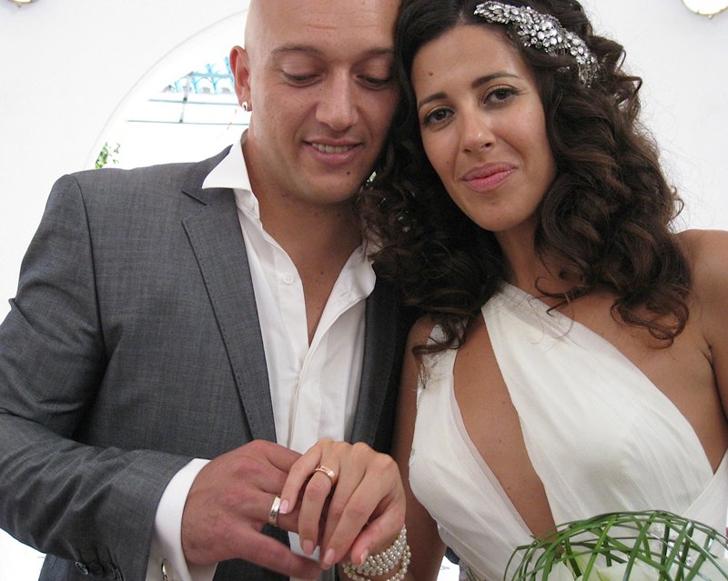 Bride and groom showing their gold wedding rings, the bride in a white gown with a jewelled hair slide and pearls