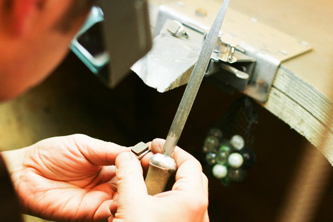 Jeweller filing a ring held in a ring clamp at the bench, a net of marbles hanging below