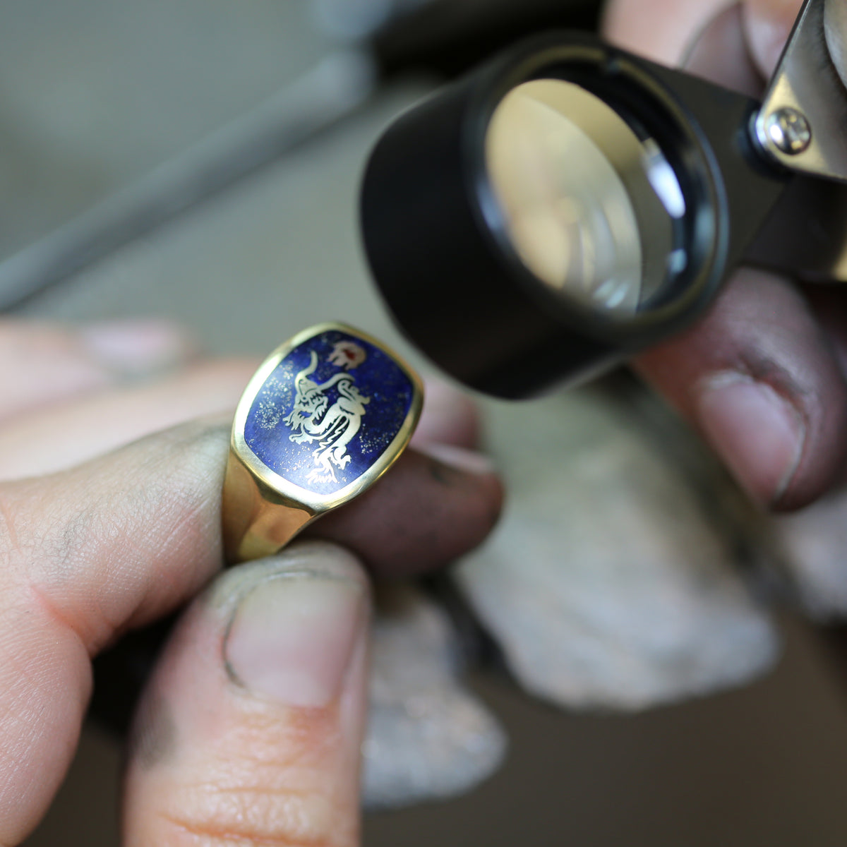 Man in a jewellery workshop is looking at a bespoke yellow gold signet ring with blue enamel and a dragon design through a magnifying glass