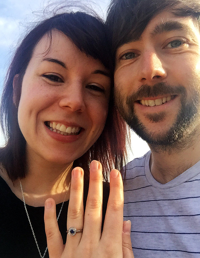 Smiling couple just engaged in Parc Güell, she holds up a ring with a dark blue stone in a halo setting