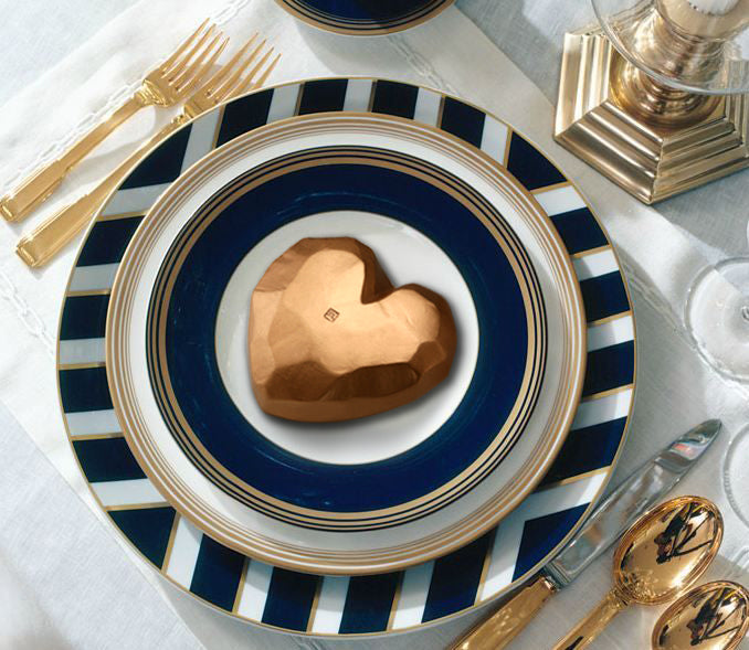 Faceted gold heart on a navy and gold striped dinner plate, laid with gold cutlery and white linen