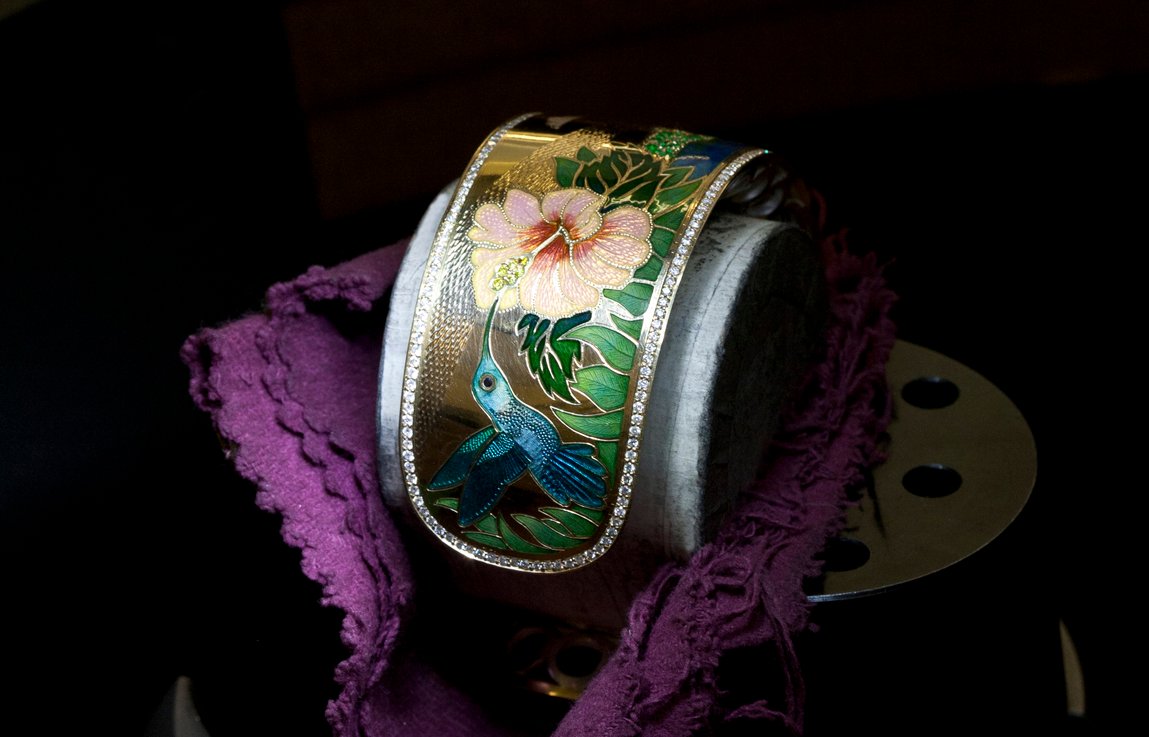 Bespoke gold cuff with an enamel hummingbird and hibiscus flower, edged with diamonds, on purple cloth