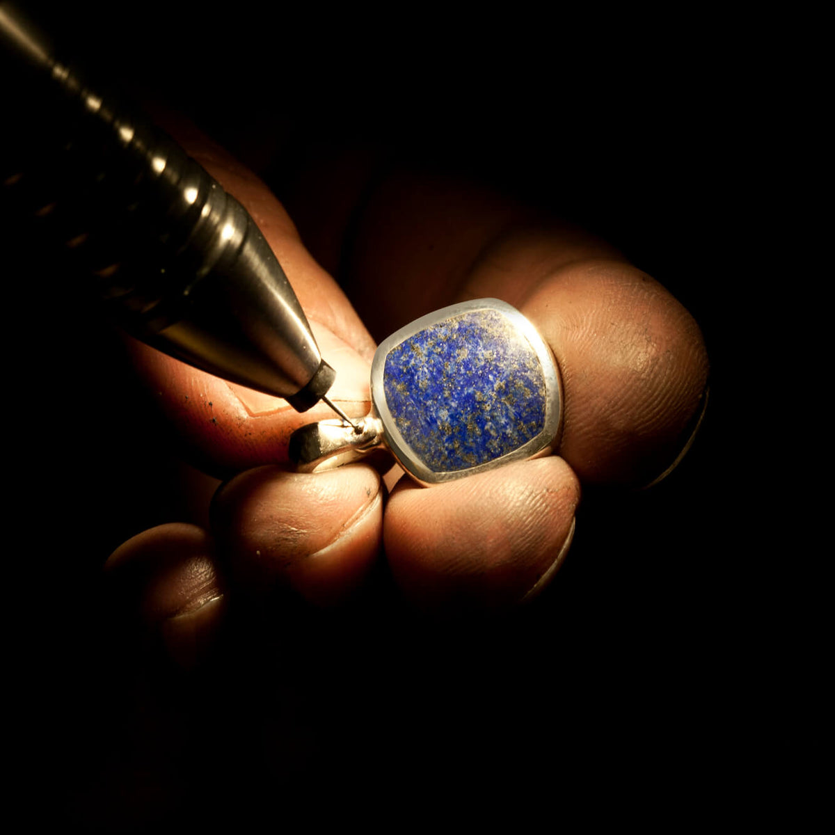 A jewellers hands in the dark working on an inlaid signet necklace with lapis lazuli