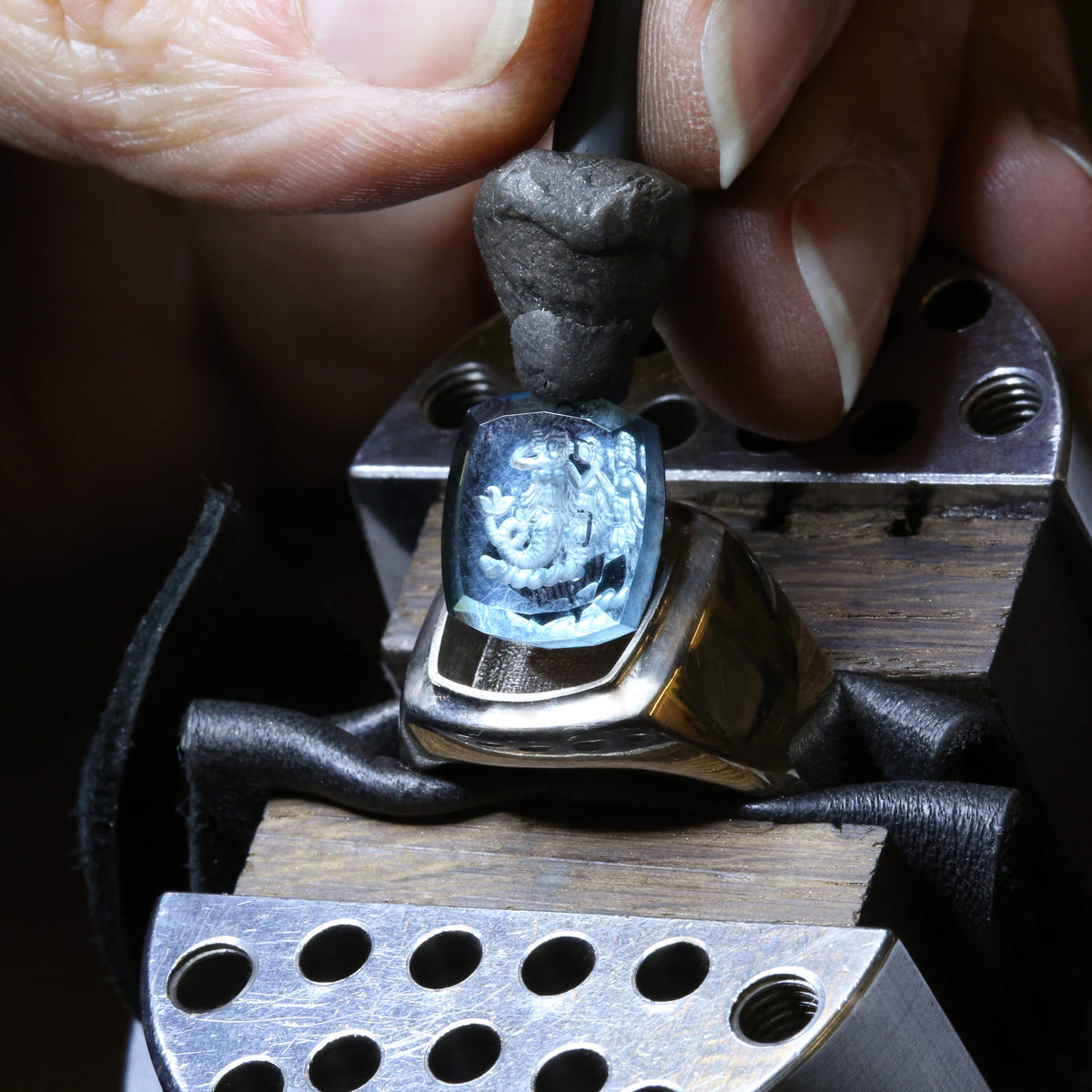 Someone in a workshop environment is setting a silver intaglio signet ring with a blue engraved stone