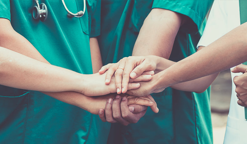 Hands of health workers in green scrubs stacked together, one wearing a gold ring, a stethoscope behind