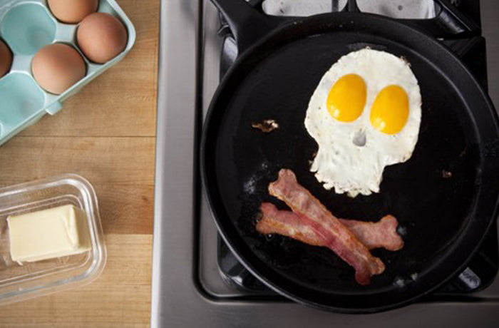Fried eggs shaped like a skull with bacon crossbones in a black frying pan on a hob