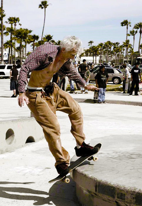 Older grey-haired man skateboarding at Venice Beach skatepark in a patterned shirt and tan trousers