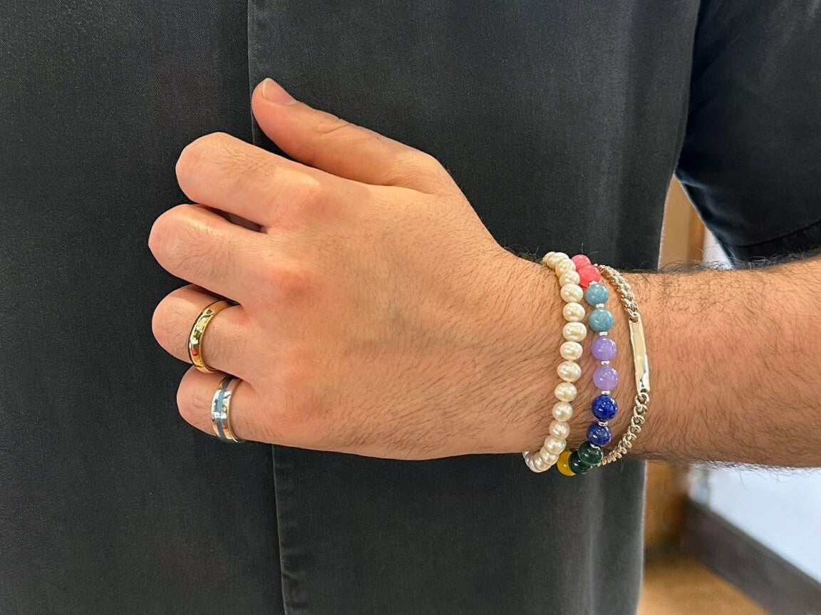 Man's wrist stacked with pearl, rainbow gemstone and silver chain bracelets, with gold and silver rings, for Pride