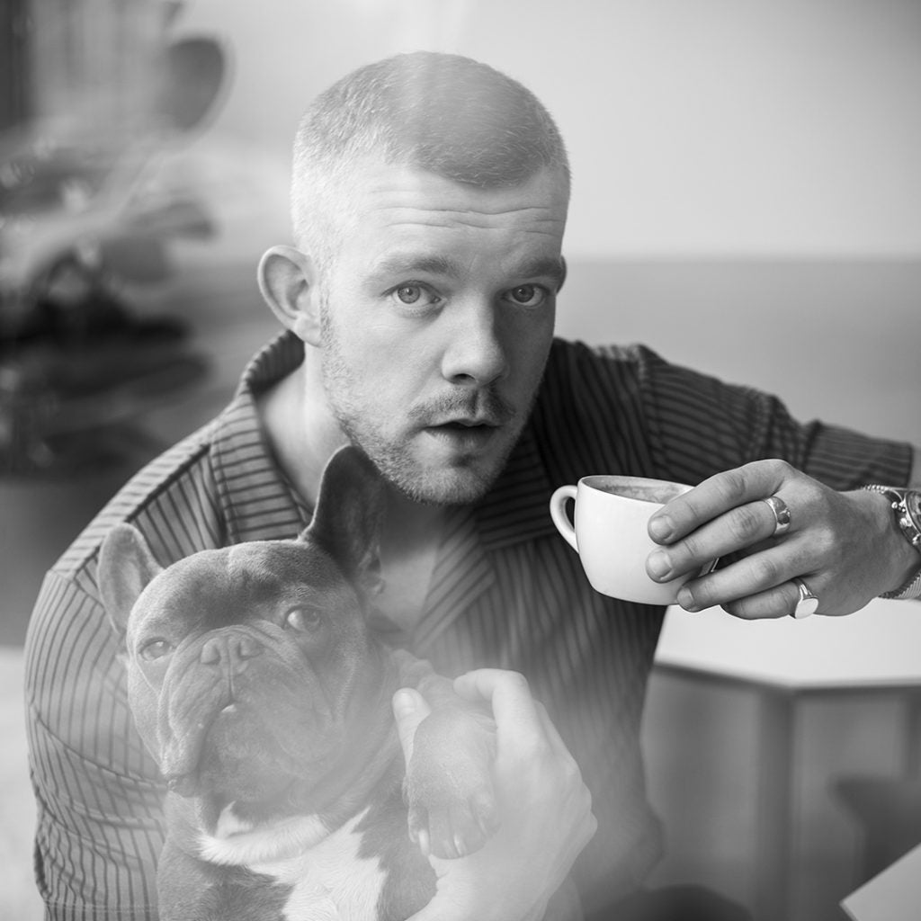 Russell Tovey in black and white holding a coffee cup and a French bulldog, a silver signet ring on his finger