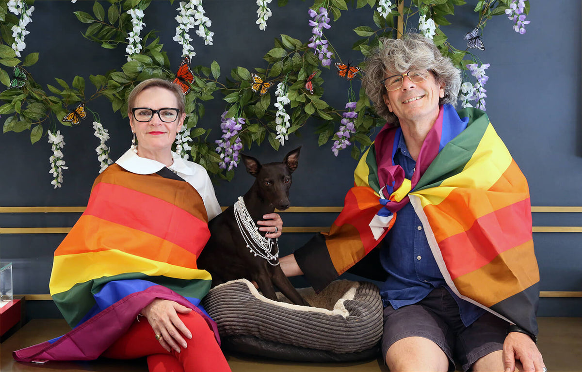 Two people wrapped in rainbow Pride flags sitting with a black dog wearing a silver chain necklace