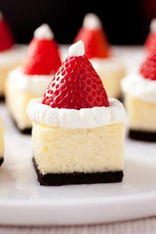 Mini cheesecakes topped with strawberries and cream piped to look like Santa hats, on a white plate
