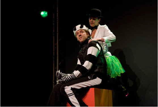 Two actors on a dark stage for Scene & Heard, one in a striped costume and one in a top hat and green tutu