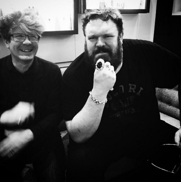 Kristian Nairn showing a large black stone ring and silver chain bracelet beside a smiling man, black and white