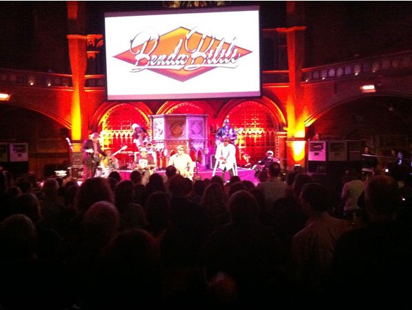 Staff Benda Bilili performing on stage in a red-lit venue, their name on a screen above the band