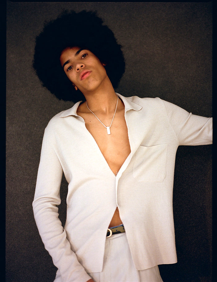 Young man with an afro in an open cream shirt wearing a silver rectangular pendant necklace