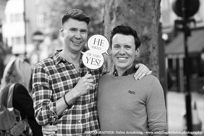 Two smiling men holding a sign reading "He asked... He said yes!" on a London street, black and white