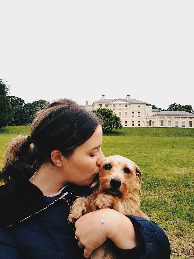 Woman kissing Otto the wire-haired dachshund in a park, a silver bracelet on her wrist and a white house behind