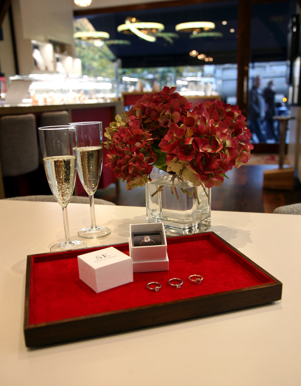 Engagement rings on a red velvet tray with a ring box, champagne and red hydrangeas in our shop