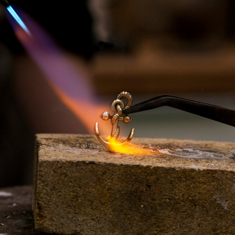 Silver anchor pendant being soldered with a torch flame on a firebrick at the jeweller's bench
