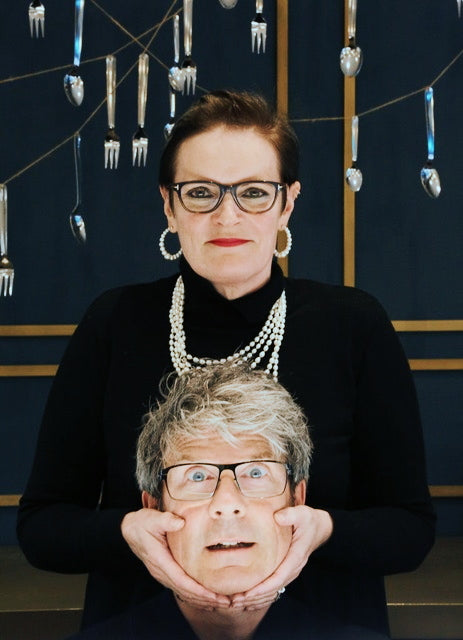 Woman in pearl necklaces and hoop earrings holding a grey-haired man's face in her hands, under hanging cutlery