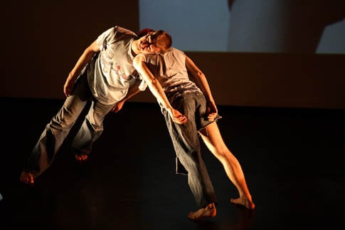 Two dancers leaning against each other on a dark stage in Sum of Parts at Sadler's Wells