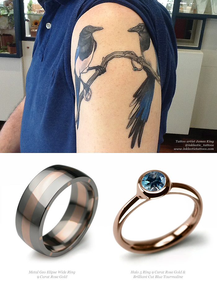 Magpie pair tattoo on a man's upper arm, above a rose gold striped band and a rose gold blue tourmaline ring