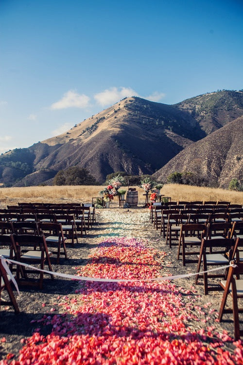 Outdoor wedding ceremony with rows of wooden chairs and a rose petal aisle leading towards brown hills