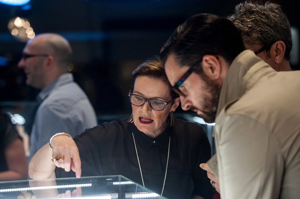 Jane Best in glasses and a long pendant pointing into a lit jewellery display case as two men look on