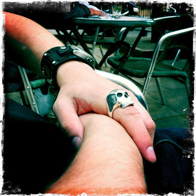 Large silver skull ring and black leather cuff worn on a man's hand at an outdoor café table