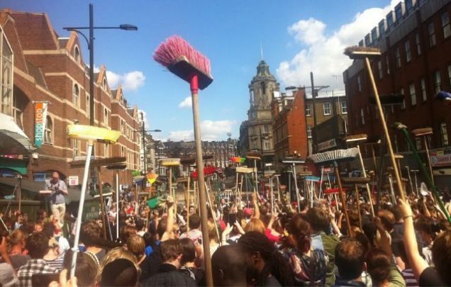 Crowd of people holding brooms aloft in a London street during the 2011 riot clean-up