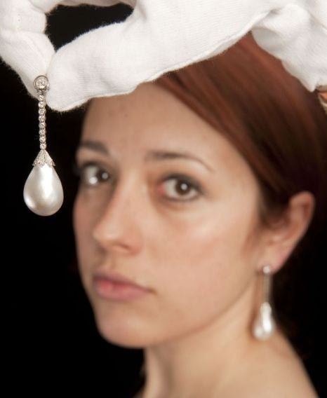 Gloved hand holding a large drop pearl and diamond earring beside a woman wearing its pair