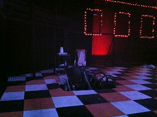 Dark theatre stage for Faust with a checkerboard floor, a wheelchair and window frames lit by red bulbs
