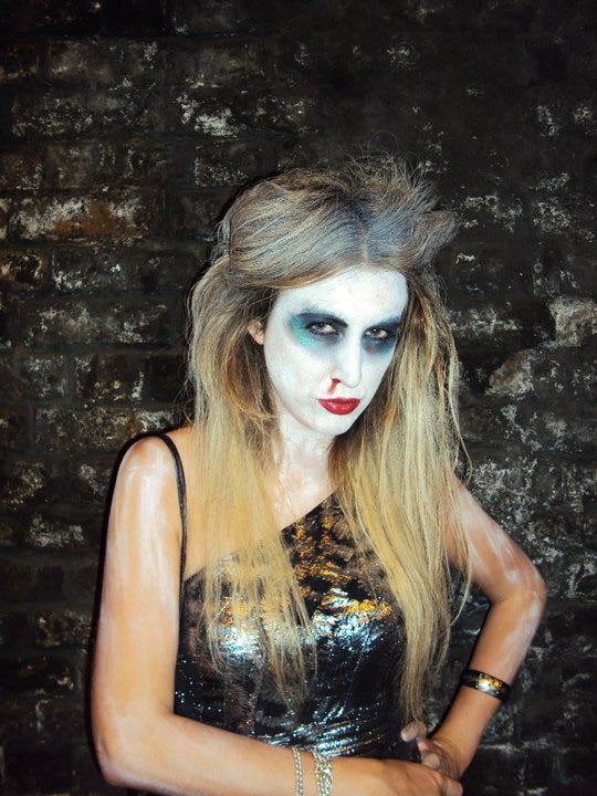 Woman in zombie make-up and a metallic dress, wearing a gold cuff and chain bracelets, against a brick wall