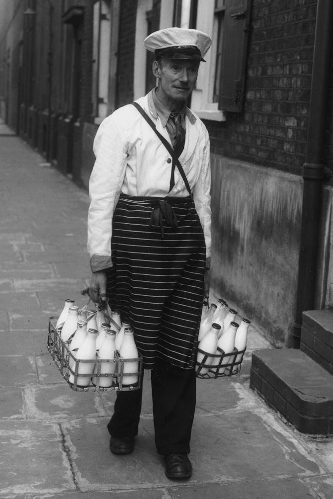 Black and white archive photograph of a milkman in cap and apron carrying crates of milk bottles