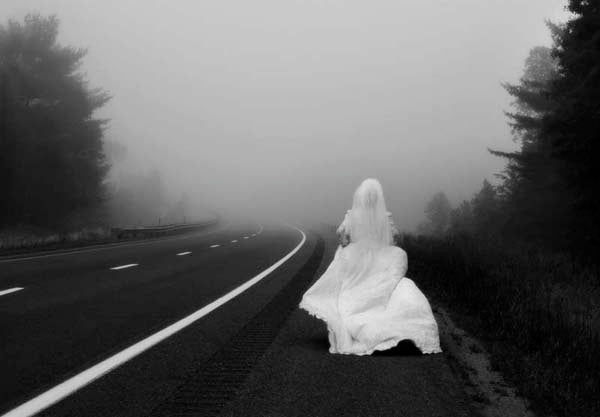 Black and white of a woman in a white wedding dress sitting at the edge of a misty road