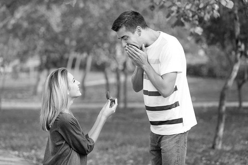 Woman kneeling in a park holding out an open ring box to a surprised man covering his mouth