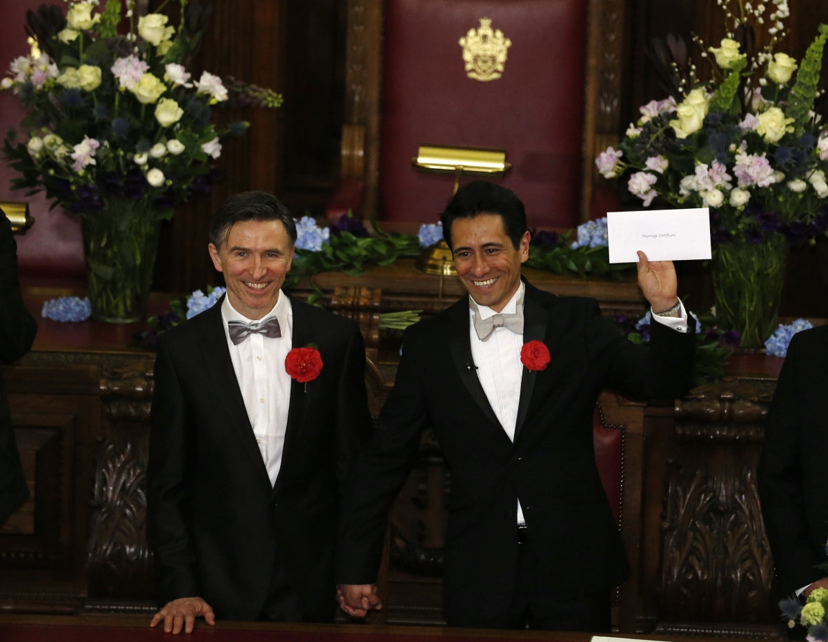Two grooms in black tuxedos with red flowers smiling at their wedding, one holding up a certificate